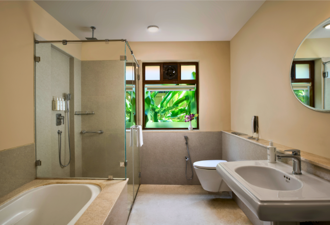Spacious bathroom with bathtub, glass rainfall shower and tropical garden views at Mercure Goa Devaaya Resort